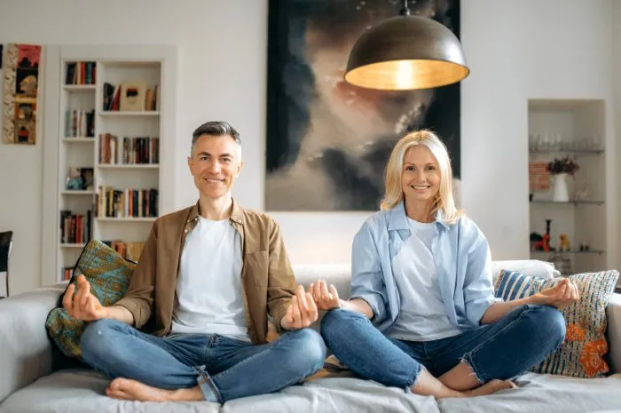 healthy lifestyle married middle aged happy couple in love, husband and wife in casual wear, doing yoga at home on sofa, sitting in lotus position at living room, meditating,looking at camera smiling