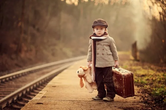 adorable boy on a railway station, waiting for the train with suitcase and teddy bear