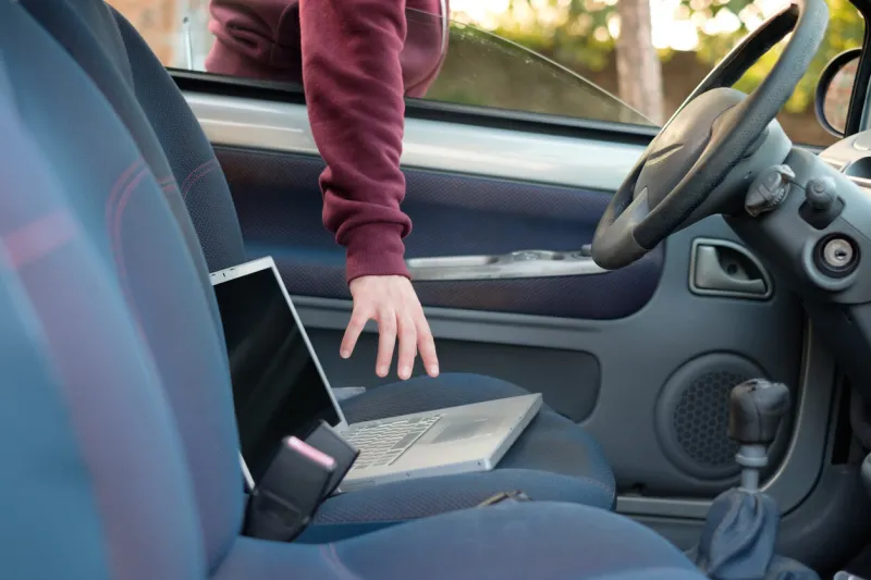 hooded thief stealing a computer laptop from a parked car