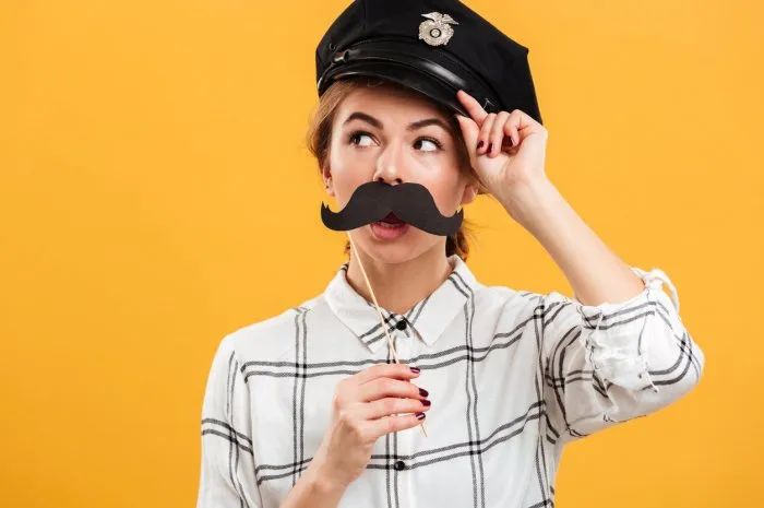 portrait of funny woman in plaid shirt and police cap holding paper moustache at her face, isolated over yellow background