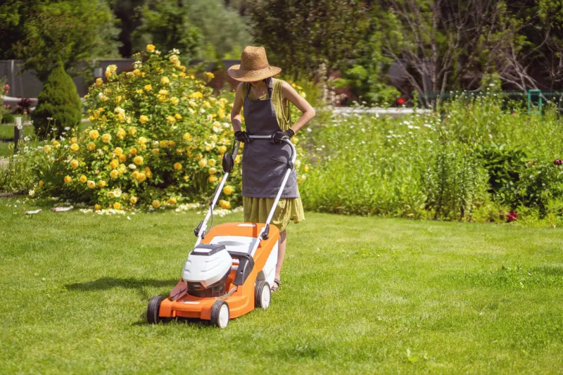 a young girl in a straw hat is mowing a lawn in the backyard with an orange lawn mower a woman gardener is trimming grass with the grass cutter a lawnmower is cutting a lawn on a summer sunny day
