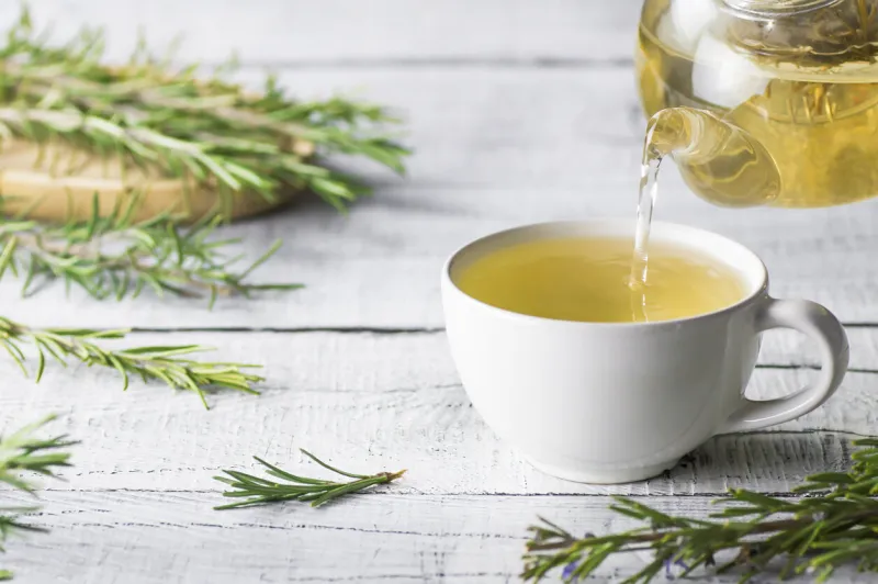 white cup of healthy rosemary tea pouring from teapot with fresh rosemary bunch on white wooden rustic background, winter herbal hot drink concept, salvia rosmarinus