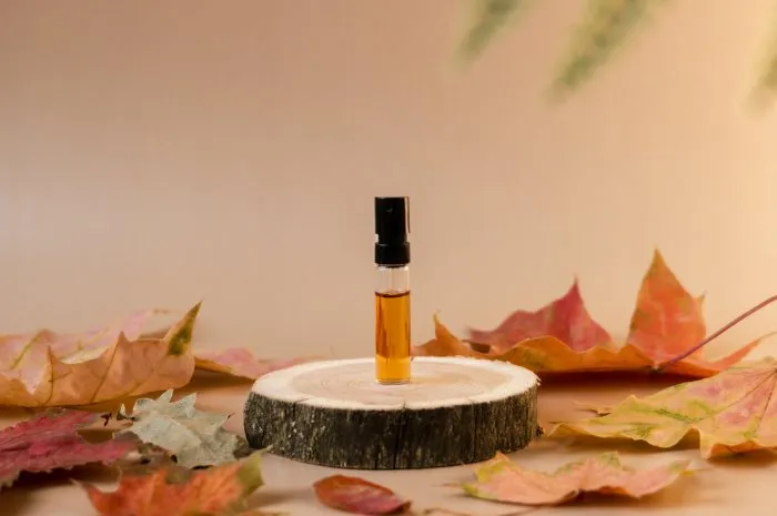 glass perfume sample with brown liquid on a wooden tray standing on beige background with fallen leaves around luxury and natural autumn cosmetics presentation tester on a woodcut front view