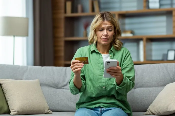 a woman sitting on sofa at home worriedly holding phone and credit card problem with the account, online payment, blocking, bankruptcy, fraud