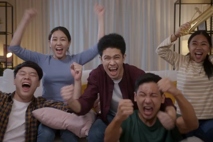 group of young adult friend man and woman asia people sit at sofa couch joy party fun game fifa world cup live tv at home chanting hand raised sign jump mad happy win exult face