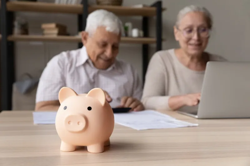 focus on piggybank on table, on background frugal elderly couple make payment using e-bank on laptop, calculates earning smile feel happy family budget control, management, investment, profit concept