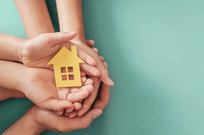 hands holding yellow paper house on blue background, family home, homeless shelter housing and home protecting insurance, mortgage concept, foster home care