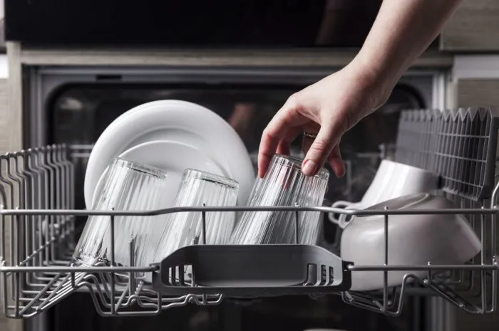 close-up of female hand loading dished to, empty out or unloading from open automatic stainless built-in dishwasher machine with clean utensils inside in modern home kitchen household domestic life