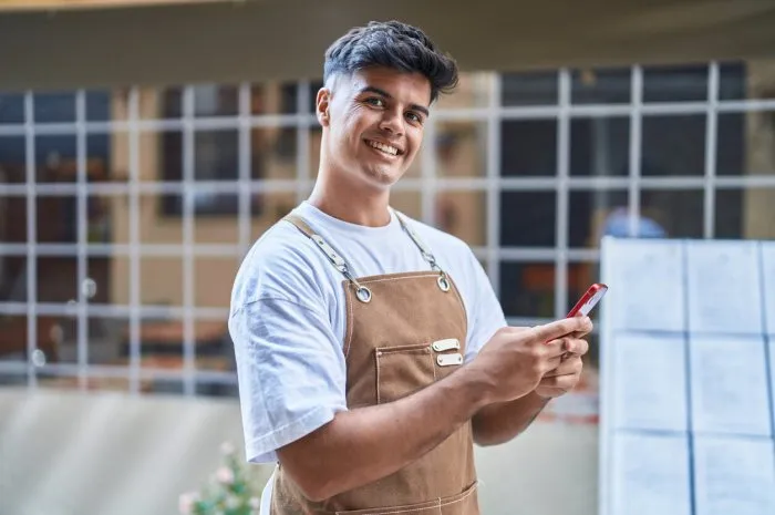 young hispanic man waiter smiling confident using smartphone at coffee shop terrace