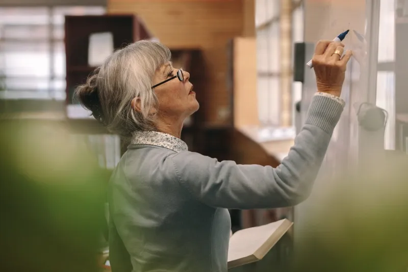 side view of a senior female teacher writing on a white board in classroom close up of a woman lecturer teaching in classroom
