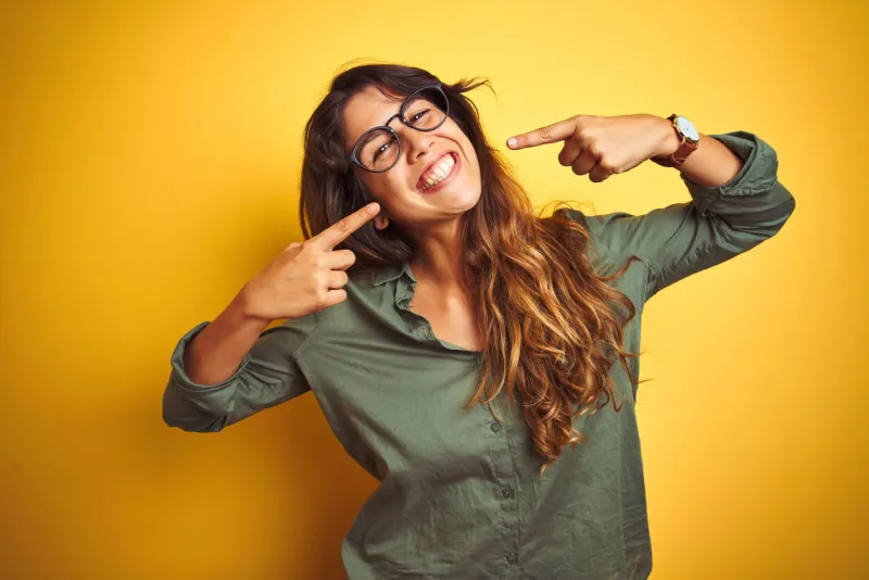 young beautiful woman wearing green shirt and glasses over yelllow isolated background smiling cheerful showing and pointing with fingers teeth and mouth dental health concept
