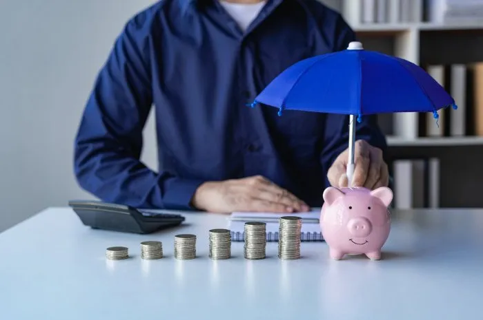 property protection concept, male hand holding open blue umbrella protecting piggy bank and house stacking gold coins on table, loan or saving money for real estate
