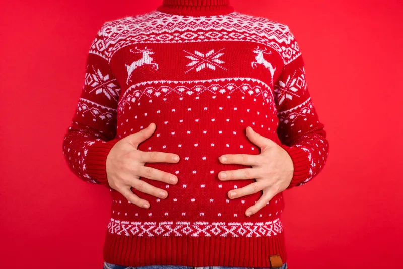 holidays overeating concept close up photo of men in red winter sweater with deers holding his big tummy isolated on red background