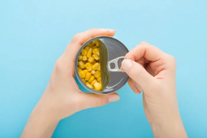 young adult woman hands holding and opening new metallic preserved can with yellow corn on light blue table background pastel color closeup point of view shot top down view