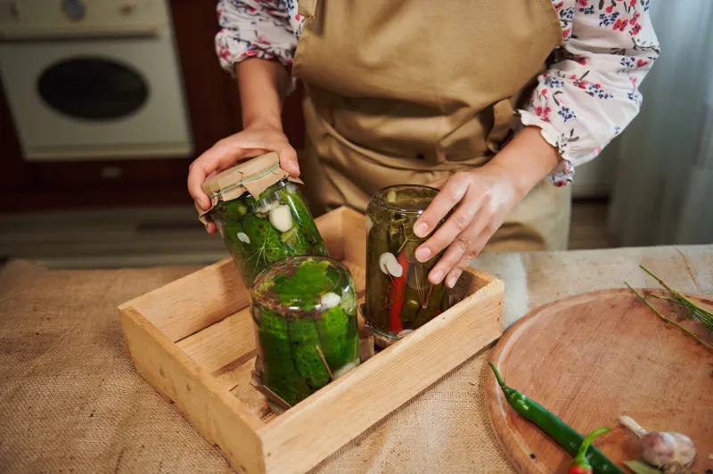 details  a housewife's hands put canned fermented pickles and pickled chili peppers with vinegar brine upside down on a wooden box in a home kitchen preparing homemade preserved for the winter