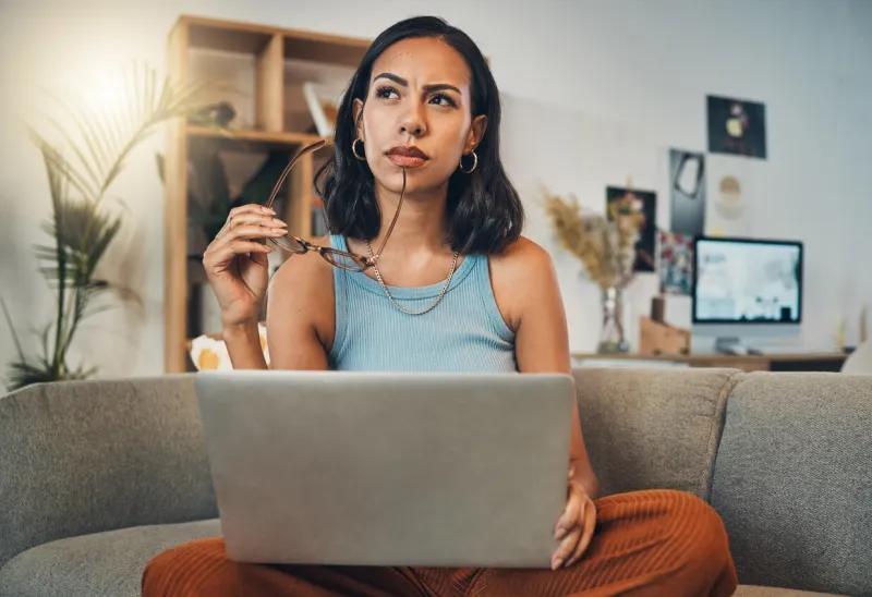 beautiful mixed race woman thinking while using laptop for blogging in living room at home hispanic entrepreneur sitting cross legged alone on lounge sofa and planning next blog post on technology