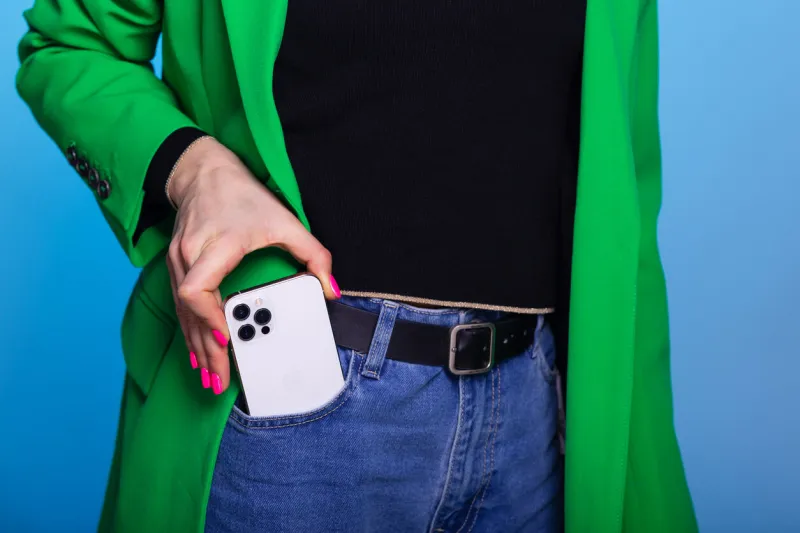 young woman takes out or puts in a pocket mobile phone woman wears green blazer and blue jeans smartphone with three cameras in front pocket on blue background mockup for print or design template