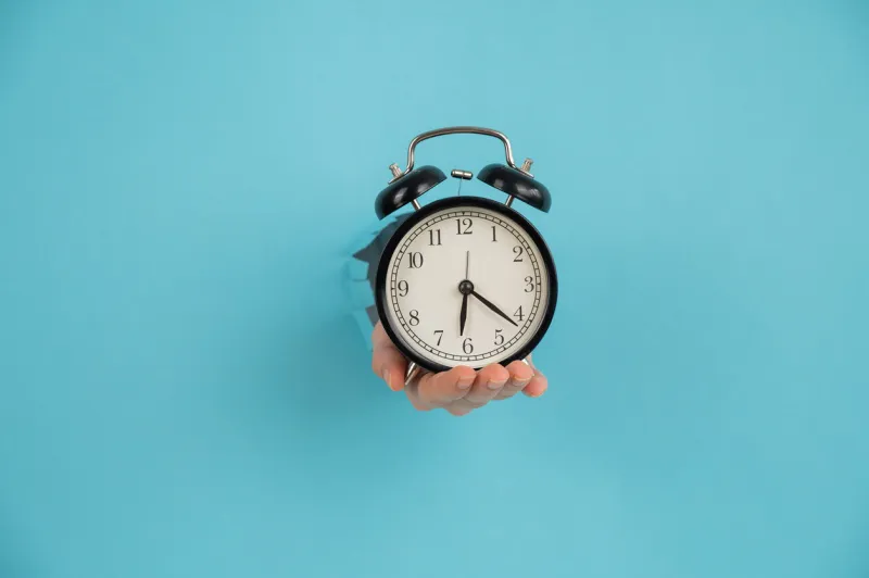 a woman's hand sticks out of a hole in a blue paper background and holds an alarm clock copy space