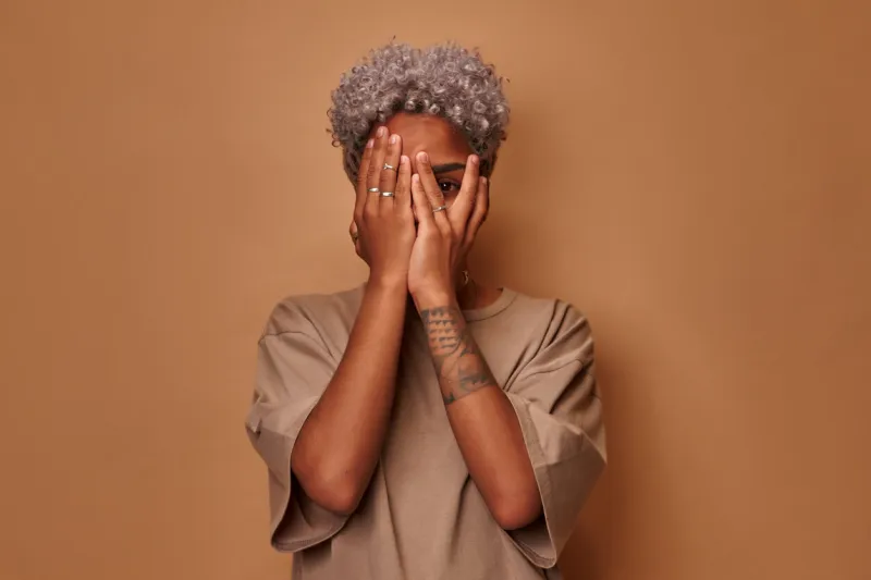 frightened dark skinned woman covers face with palms peeks through fingers sees something horrible and unexpected, hides from all difficulties intrigued what happening, against studio beige background