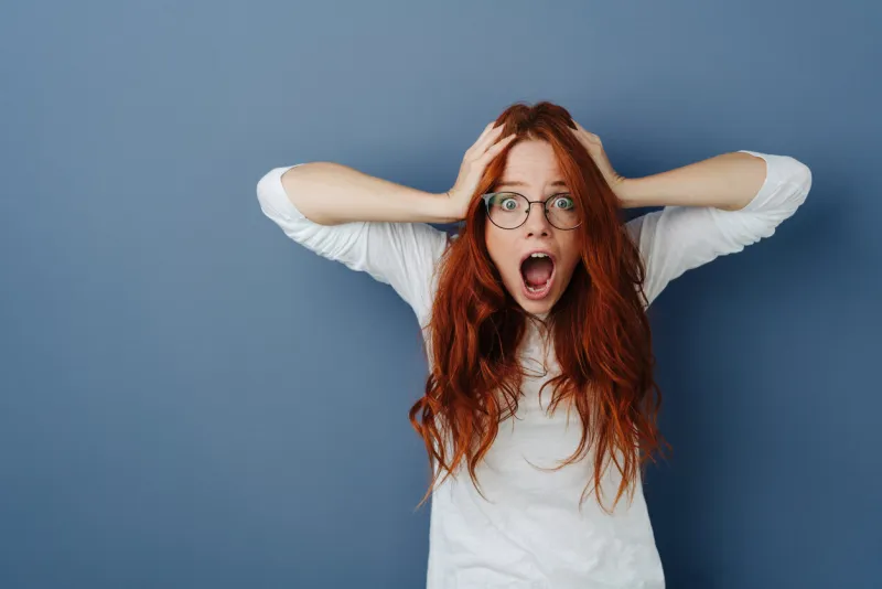 aghast young woman in a panic staring at the camera with mouth open, wide eyes and hands to her long red hair over a blue studio background with copy space
