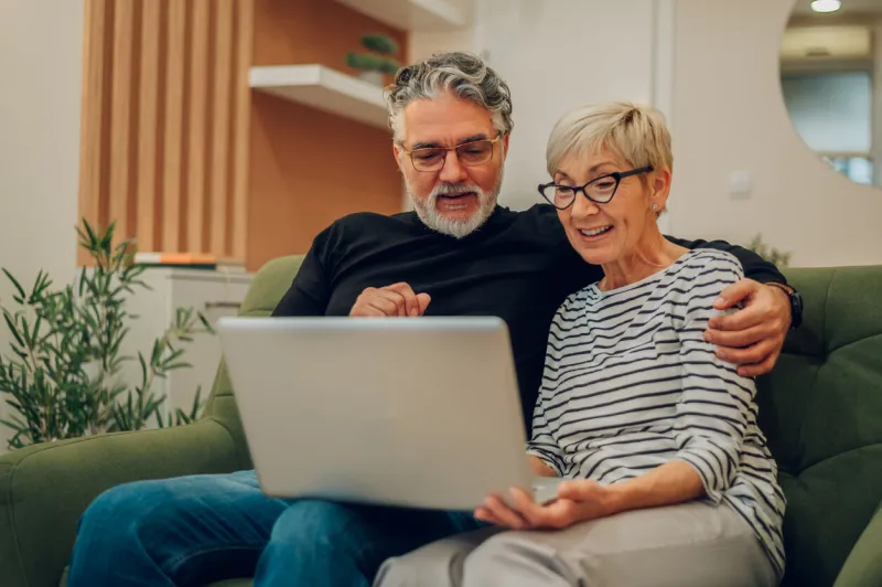 portrait of a senior married happy couple embracing using laptop together and sitting on a couch at home smiling elderly spouses reading news, shopping online older people and computer concept