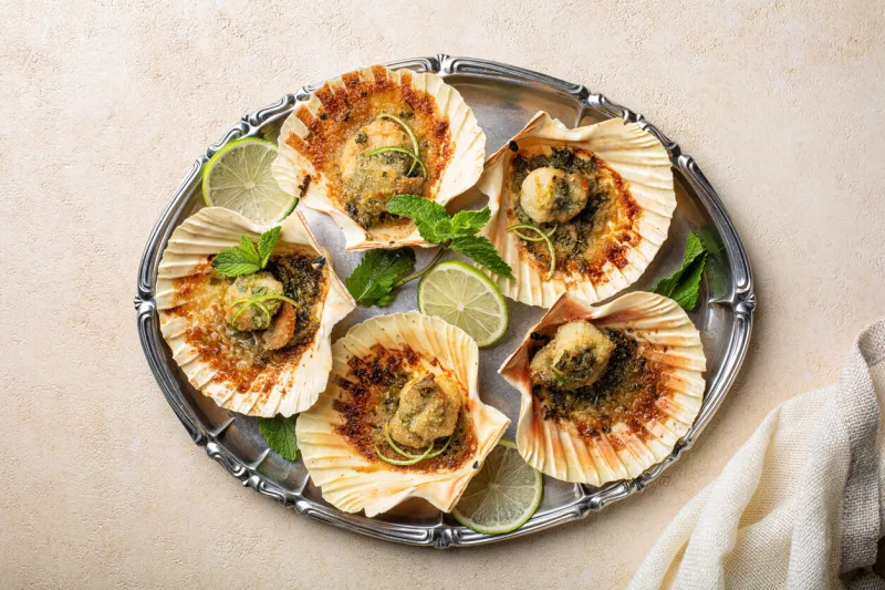top view of baked scallops with breadcrumbs, lime, parsley and mint light beige background