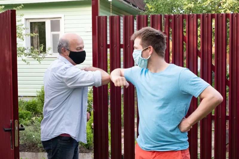 two men in medical mask greeting each other with elbows on sunny day near red fence