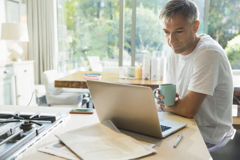 man drinking coffee and working at laptop in kitchen