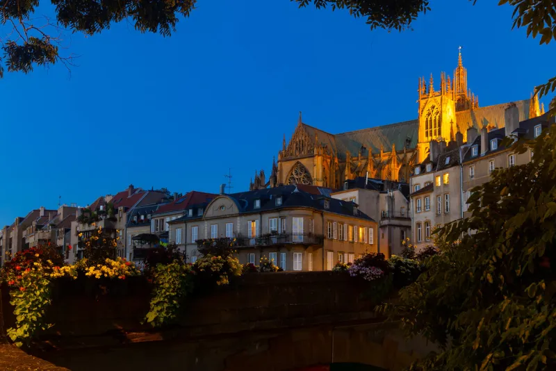 scenic nighttime view of summer metz cityscape overlooking illuminated majestic gothic roman catholic cathedral towering over residential buildings on waterfront along moselle river, france