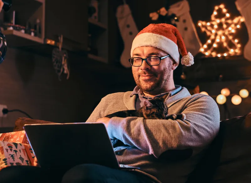 man in santa hat and glasses dressed in warm sweater holds cat and using laptop celebrating christmas eve at home