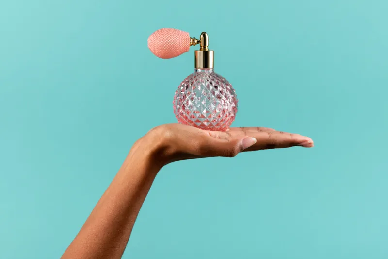crop unrecognizable african american female demonstrating pink crystal vintage bottle with spray of perfume on turquoise background