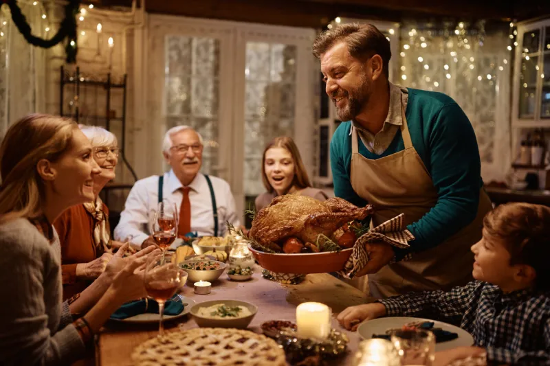 happy man bringing roast turkey at the table during thanksgiving dinner with his multigeneration family