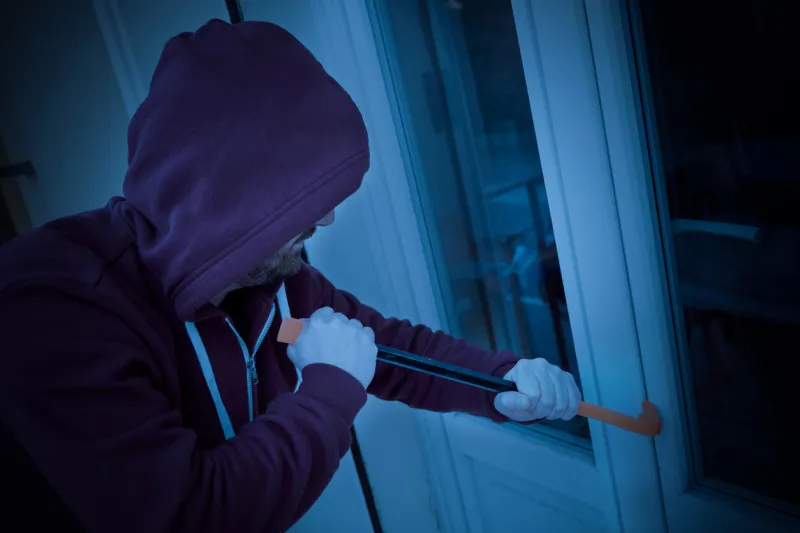 hooded burglar forcing window lock to make a theft in a house at night