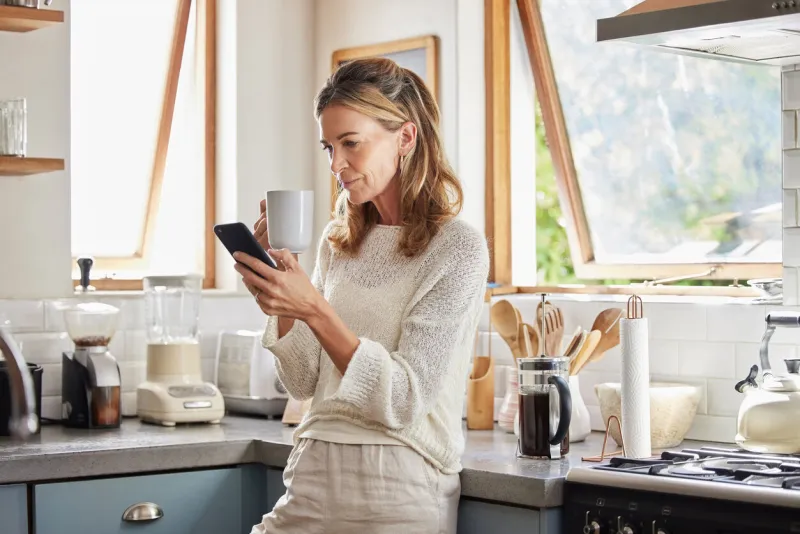 coffee, phone and senior woman in kitchen browsing social media, text message or online app at home relax, tea and reading news or mobile online web surfing with female from canada on 5g smartphone