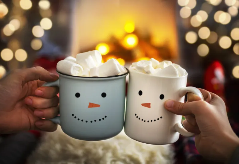 mom and child are relaxing together on a cozy winter evening by the fireplace, close-up of two hands with snowman face cup of hot cocoa with marshmallows christmas holidays, happy moments at home
