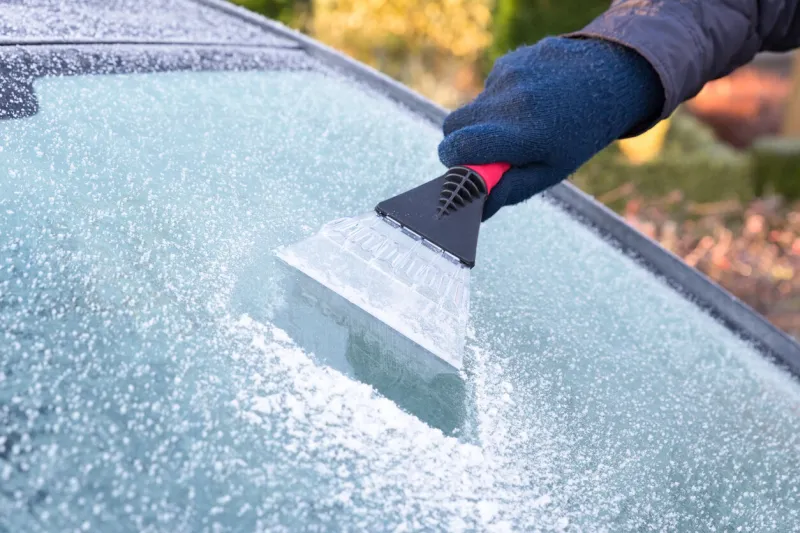 hand wearing blue glove scratching ice from car window the ice scraper is an extension of the arm part of the windshield is ice free only part of the window is visible symbol or concept of winter season, cold, frost, weather, sight