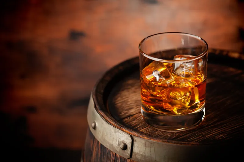 glass of whiskey with ice cubes on the old barrel with copy space on wooden background