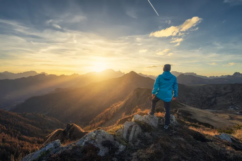 man on stone on the hill and beautiful mountains in haze at colorful sunset in autumn dolomites, italy sporty guy, mountain ridges in fog, orange grass and trees, blue sky with sun in fall hiking