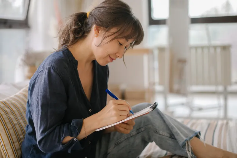 asian young smiling woman making notes in notebook, working at home on bed in cozy bedroom concept of freelancer, remote work, home office