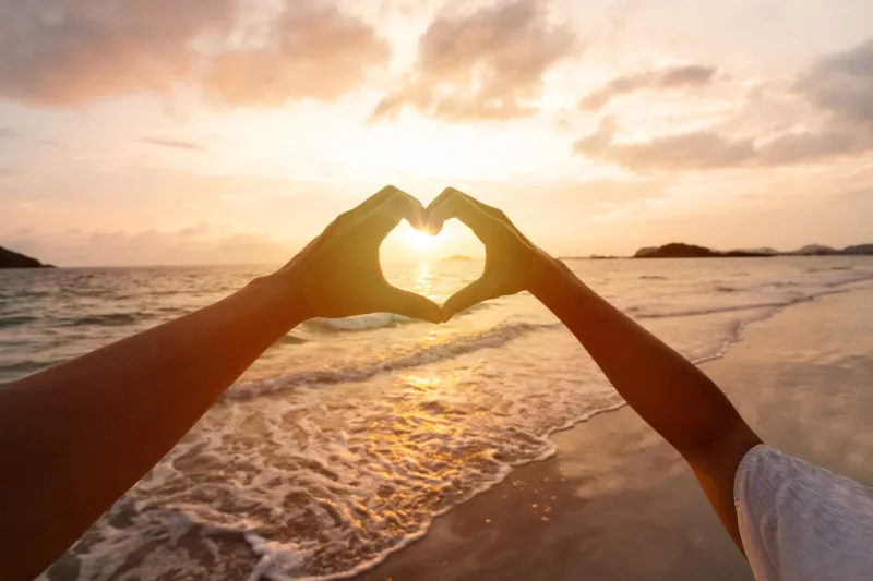 young couple traveler making heart shape with hands on the beach at sunset, lovers on honeymoon and valentine's day concept
