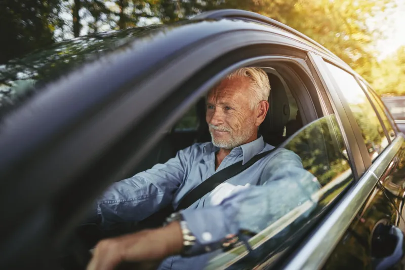senior man smiling while sitting alone in his car driving along a tree lined road in the country