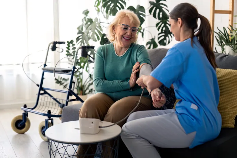 beautiful senior woman measuring blood pressure with help of home nurse, sitting on sofa in living room, feeling satisfied and happy with home care female nurse assisting senior woman to measure blood pressure