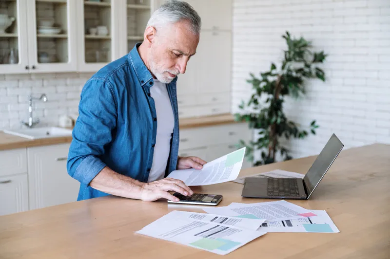 focused middle aged man making calculations of utility payment holding paper bill invoice in hand at table serious businessman doing paperwork for paying taxes online using laptop at home