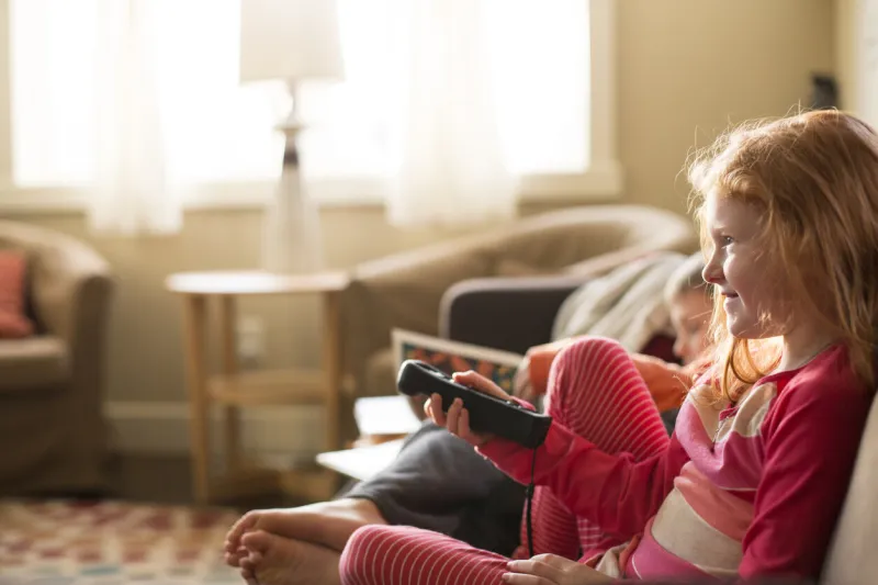 young, redheaded girl wearing a colorful pink and coral striped top and pants holds television remote in a sunny, bright living room and smiles at what she is watching on tv her brother is in the background