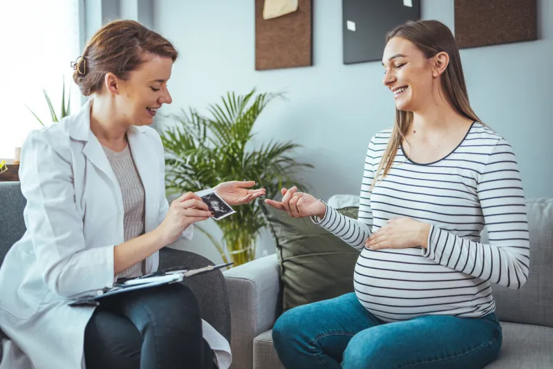 young woman smiles while looking at her baby's ultrasound image she is meeting with a home healthcare nurse pregnant woman and doctor looking at her baby ultrasound picture