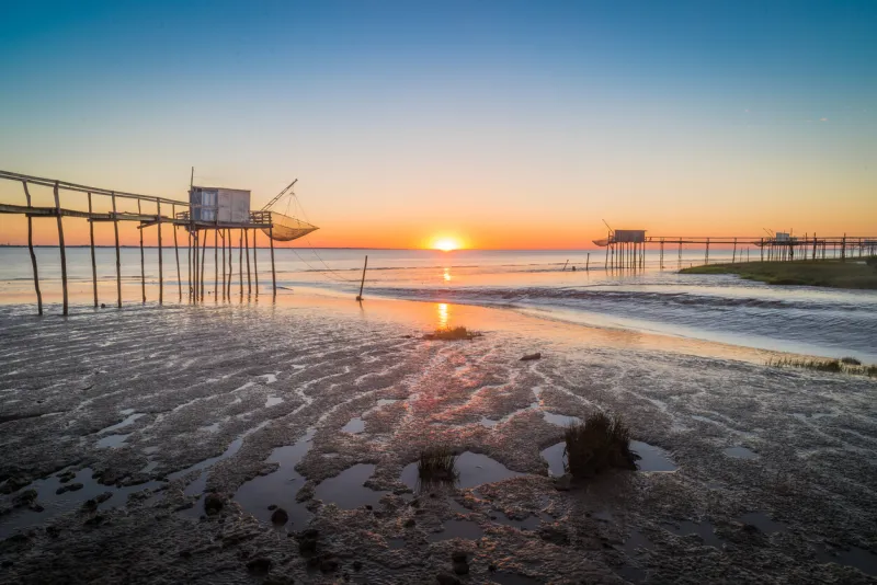sun setting on horizon with traditional fishing huts on shores at low tide in charente maritime, france, near la rochelle