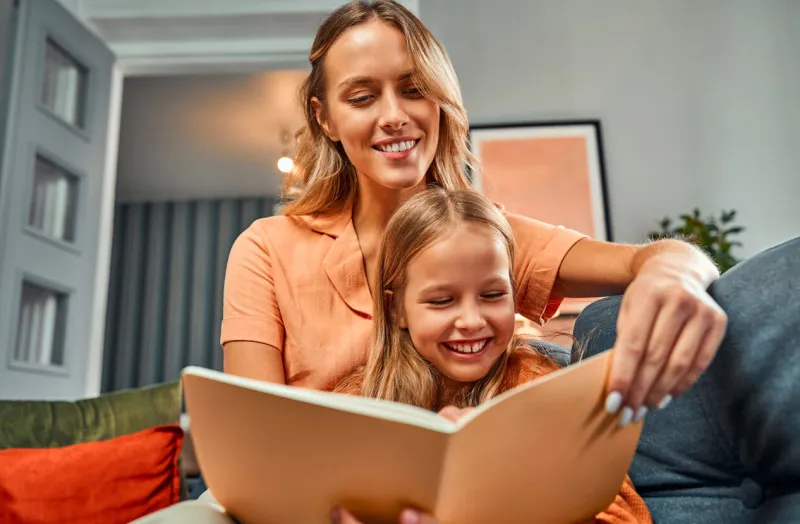 excitement from learning young mother enjoying spending time with child while doing homework cute little girl laughing while sitting near mom and reading book together at comfy sofa