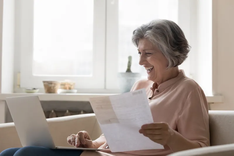 cheerful senior retired woman paying bills on internet, counting budget, using online app on laptop at home, typing, holding paper document, invoice, receipt, smiling, laughing