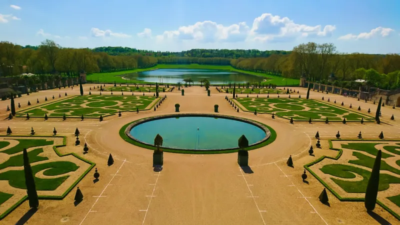 estate of versailles, paris, france - april 20, 2018  aerial view of a designed garden in versailles with trees, grass, a fountain and a lake in patterns under bright blue sky and clouds