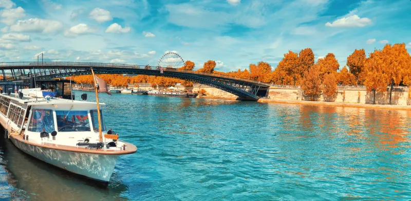 paris, seine river in autumn passenger ship in front of pedestrian bridge passerelle leoopold-sedar-senghor and autumn leaves on the river bank panoramic toned image